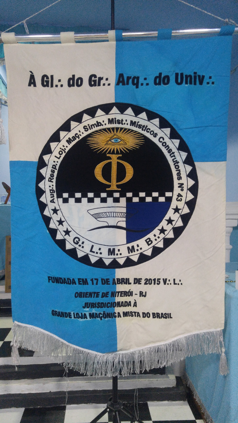 Estandarte da Loja Místicos Construtores nº 43: quadrantes azul-claro e branco, emblema circular com orla denteada, Delta radiante com o Olho que Tudo Vê, a letra Phi, faixa quadriculada e a silhueta do MAC de Niterói, com franja e borlas prateadas.